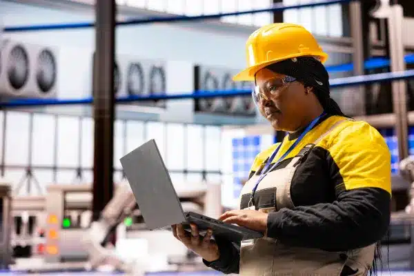 Technician with a laptop managing IoT systems in an industrial environment, wearing safety gear.