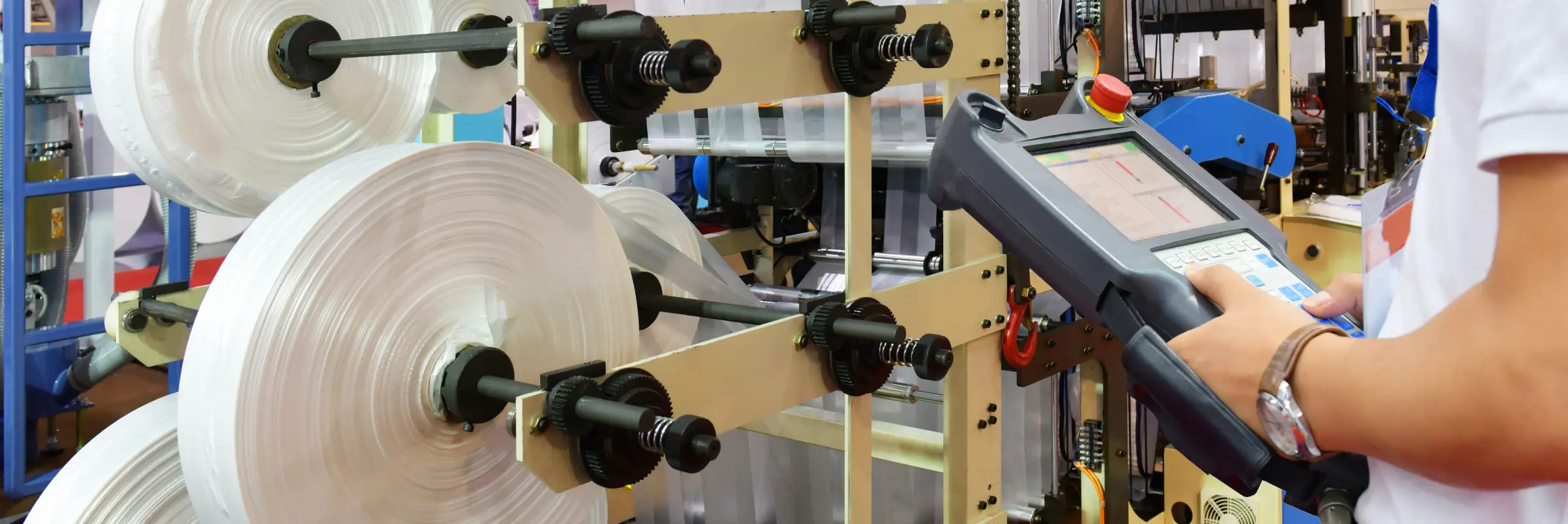 Worker operating a handheld digital control panel next to large rolls of plastic film on industrial manufacturing equipment