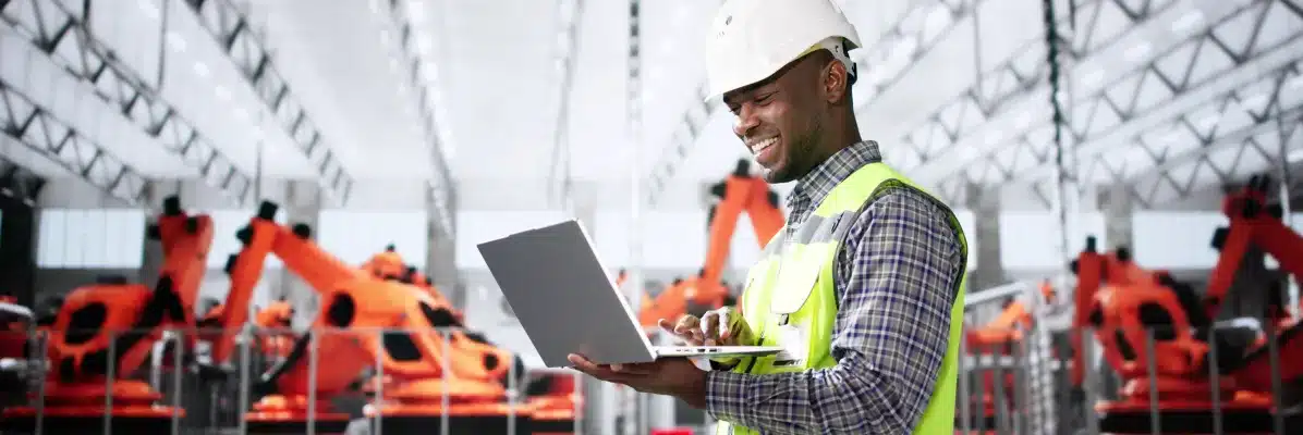 Male engineer using laptop for IIoT monitoring architecture in automated robotic manufacturing facility.